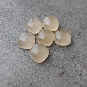 May include: Six translucent, rounded square-shaped stones with a pale yellow hue are arranged on a gray surface. Each stone has a smooth, polished appearance, reflecting light. The stones are similar in size and shape, suggesting they are part of a set or collection.