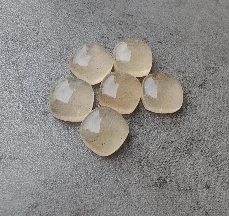 May include: Six translucent, rounded square-shaped stones with a pale yellow hue are arranged on a gray surface. Each stone has a smooth, polished appearance, reflecting light. The stones are similar in size and shape, suggesting they are part of a set or collection.