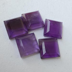 Peut inclure: Cinq pierres pr&eacute;cieuses d'am&eacute;thyste taill&eacute;es en carr&eacute;. Chaque gemme pr&eacute;sente une teinte violette profonde avec des bords facett&eacute;s, captant la lumi&egrave;re. Les pierres sont dispos&eacute;es sur une surface claire, mettant en valeur leur couleur et leur forme.