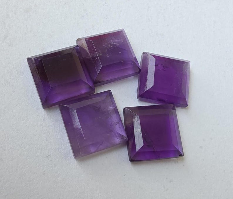 Peut inclure: Cinq pierres pr&eacute;cieuses d'am&eacute;thyste taill&eacute;es en carr&eacute;. Chaque gemme pr&eacute;sente une teinte violette profonde avec des bords facett&eacute;s, captant la lumi&egrave;re. Les pierres sont dispos&eacute;es sur une surface claire, mettant en valeur leur couleur et leur forme.