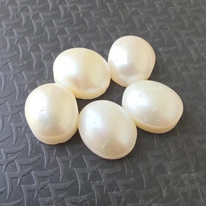 Op de afbeelding: Vijf cr&egrave;mekleurige, ovale parels met een parelmoerglans. De parels zijn gerangschikt op een donkergrijze, getextureerde ondergrond, waardoor een contrast ontstaat dat hun lichtgevende kwaliteit benadrukt. De parels zijn glad en hebben een natuurlijk, organisch uiterlijk.