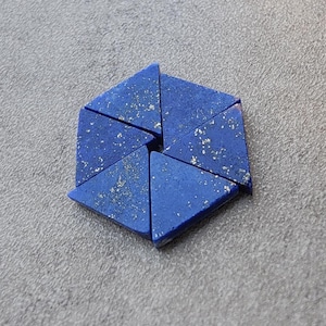 May include: A collection of six triangular, deep blue lapis lazuli stones arranged in a hexagonal shape. The stones have flecks of gold throughout, and are set against a gray background.