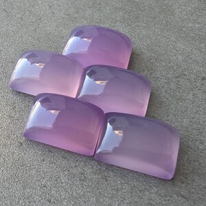 Pu&ograve; includere: Cinque pietre preziose rettangolari color lavanda lucide sono disposte su una superficie grigia. Le pietre hanno bordi arrotondati e una finitura lucida, che riflette la luce. Le pietre preziose sono di un viola morbido e traslucido.