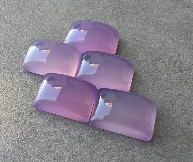 Pu&ograve; includere: Cinque pietre preziose rettangolari color lavanda lucide sono disposte su una superficie grigia. Le pietre hanno bordi arrotondati e una finitura lucida, che riflette la luce. Le pietre preziose sono di un viola morbido e traslucido.