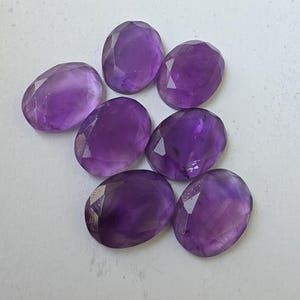 May include: Seven faceted, oval-shaped amethyst gemstones in varying shades of purple are arranged on a white surface. The gemstones have a polished, reflective surface, with visible facets that catch the light.