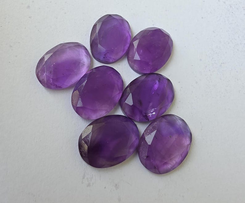 May include: Seven faceted, oval-shaped amethyst gemstones in varying shades of purple are arranged on a white surface. The gemstones have a polished, reflective surface, with visible facets that catch the light.