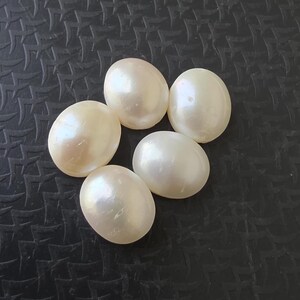 Op de afbeelding: Vijf ovale parels in een cr&egrave;mewitte kleur liggen op een donkere, getextureerde ondergrond. De parels hebben een gladde, iriserende glans die het licht reflecteert. Deze edelstenen zijn ideaal voor het maken van sieraden.