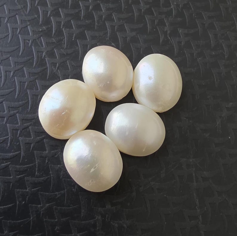 Op de afbeelding: Vijf ovale parels in een cr&egrave;mewitte kleur liggen op een donkere, getextureerde ondergrond. De parels hebben een gladde, iriserende glans die het licht reflecteert. Deze edelstenen zijn ideaal voor het maken van sieraden.