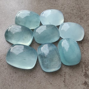 Peut inclure: Huit pierres précieuses d'aigue-marine lisses, de couleur bleu clair, chacune ayant une forme et une taille légèrement différentes, sont disposées en cercle sur une surface grise.