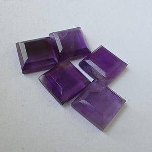 Peut inclure: Cinq pierres pr&eacute;cieuses d'am&eacute;thyste taill&eacute;es en carr&eacute;, de diff&eacute;rentes nuances de violet. Les pierres sont facett&eacute;es, avec des angles et des plans visibles, et sont dispos&eacute;es sur une surface blanche. Les pierres ont une surface polie et r&eacute;fl&eacute;chissante.