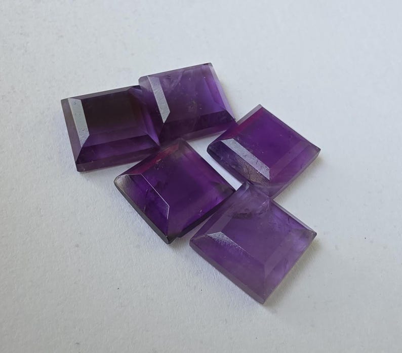 Peut inclure: Cinq pierres pr&eacute;cieuses d'am&eacute;thyste taill&eacute;es en carr&eacute;, de diff&eacute;rentes nuances de violet. Les pierres sont facett&eacute;es, avec des angles et des plans visibles, et sont dispos&eacute;es sur une surface blanche. Les pierres ont une surface polie et r&eacute;fl&eacute;chissante.