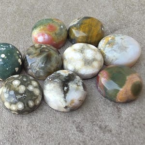 May include: A collection of nine polished stones in various colors, including green, brown, white, and red. The stones have a smooth, rounded shape and are likely used for decorative or metaphysical purposes.
