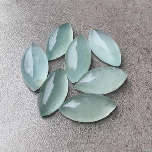 Op de afbeelding: Zeven lichtblauwgroene marquise-geslepen edelstenen op een grijze ondergrond. De gepolijste edelstenen hebben een glanzende afwerking en hun langwerpige vorm doet denken aan een blad of een oog. De doorschijnende stenen laten licht door.