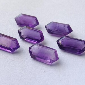 K&ouml;nnte beinhalten: Sechs facettierte Amethyst-Edelsteine mit sechseckiger Form liegen auf einer wei&szlig;en Oberfl&auml;che. Die Edelsteine sind tiefviolett und haben eine polierte, reflektierende Oberfl&auml;che. Geeignet f&uuml;r die Schmuckherstellung.