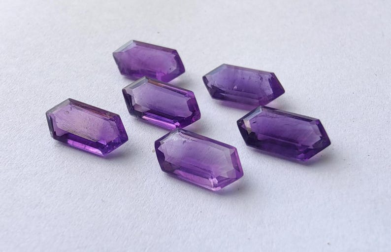 K&ouml;nnte beinhalten: Sechs facettierte Amethyst-Edelsteine mit sechseckiger Form liegen auf einer wei&szlig;en Oberfl&auml;che. Die Edelsteine sind tiefviolett und haben eine polierte, reflektierende Oberfl&auml;che. Geeignet f&uuml;r die Schmuckherstellung.