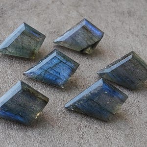 May include: Six trapezoid-shaped labradorite gemstones. The stones exhibit a play of blue, grey, and green colours, with internal patterns visible. The gemstones are displayed on a neutral surface, highlighting their polished facets.