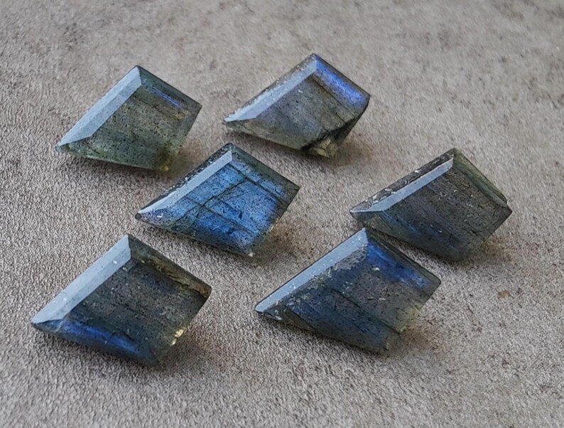 May include: Six trapezoid-shaped labradorite gemstones. The stones exhibit a play of blue, grey, and green colours, with internal patterns visible. The gemstones are displayed on a neutral surface, highlighting their polished facets.
