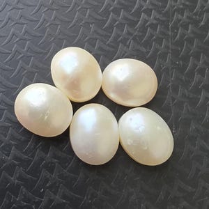 Op de afbeelding: Vijf cr&egrave;mekleurige, ovale parels met een parelmoerglans liggen op een donker, getextureerd oppervlak. De parels hebben een glad oppervlak. Geschikt voor het maken van sieraden.