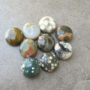 May include: A collection of nine polished, round gemstones in various colors, including green, brown, white, and speckled patterns. The stones are arranged on a gray surface, showcasing their natural textures and hues. These stones are ideal for jewelry making or collecting.