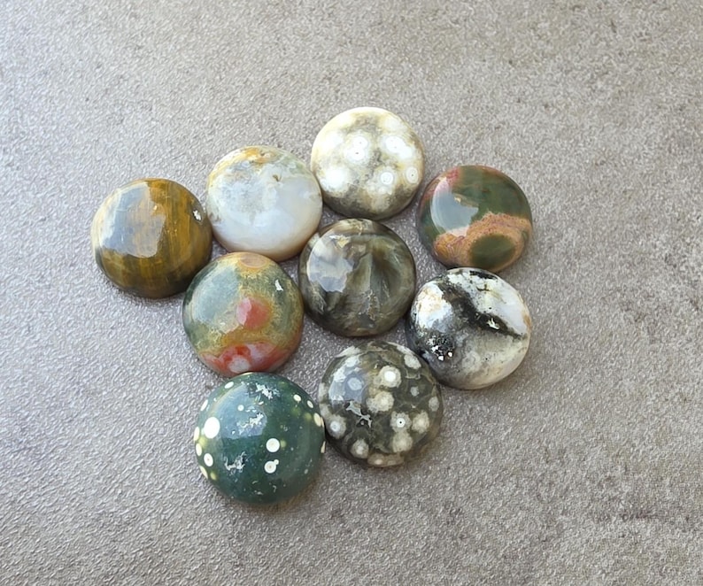 May include: A collection of nine polished, round gemstones in various colors, including green, brown, white, and speckled patterns. The stones are arranged on a gray surface, showcasing their natural textures and hues. These stones are ideal for jewelry making or collecting.