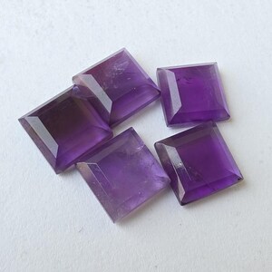 Peut inclure: Cinq pierres pr&eacute;cieuses d'am&eacute;thyste taill&eacute;es en carr&eacute;. Les pierres sont d'un violet profond avec des bords facett&eacute;s, dispos&eacute;es sur une surface blanche. Ces pierres sont probablement destin&eacute;es &agrave; la joaillerie ou &agrave; la collection.