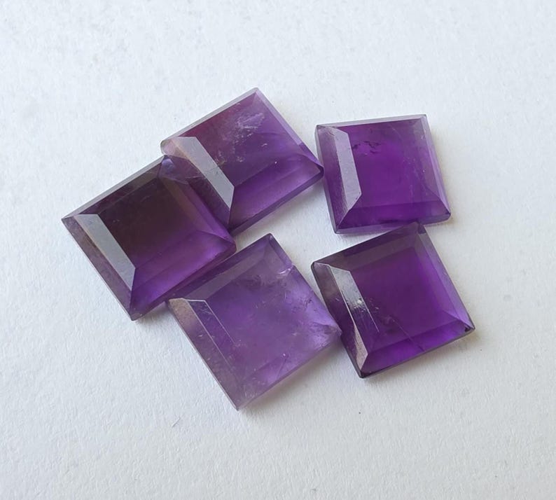 Peut inclure: Cinq pierres pr&eacute;cieuses d'am&eacute;thyste taill&eacute;es en carr&eacute;. Les pierres sont d'un violet profond avec des bords facett&eacute;s, dispos&eacute;es sur une surface blanche. Ces pierres sont probablement destin&eacute;es &agrave; la joaillerie ou &agrave; la collection.