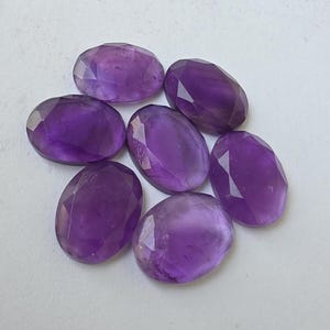 May include: Seven oval-shaped, faceted amethyst gemstones. The gems are a deep purple color and have a polished, reflective surface. The stones are arranged in a circular pattern against a white background.