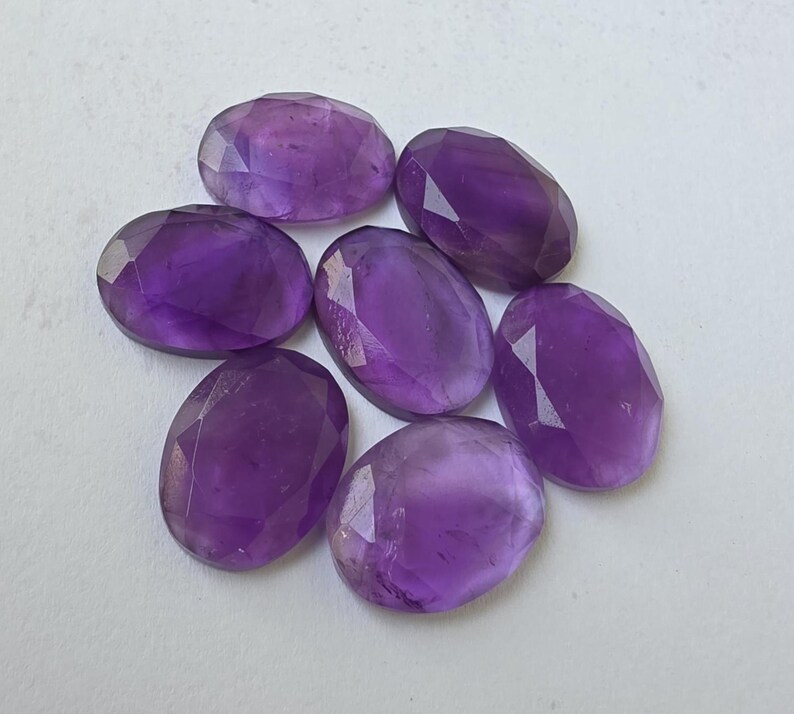 May include: Seven oval-shaped, faceted amethyst gemstones. The gems are a deep purple color and have a polished, reflective surface. The stones are arranged in a circular pattern against a white background.