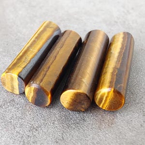 May include: Four smooth, cylindrical tiger's eye stones in shades of brown and gold. The stones are polished and have a natural, striated pattern.