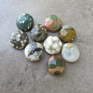 May include: A collection of nine polished gemstones in various colors, including green, brown, white, and yellow. The stones are round and have unique patterns and textures, suitable for jewelry making or collecting.