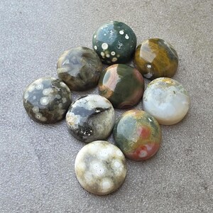 May include: A collection of nine polished gemstones in various colors and patterns. The stones include shades of green, brown, white, and red, with speckled and marbled textures. The stones are oval and round, and are arranged on a gray surface.