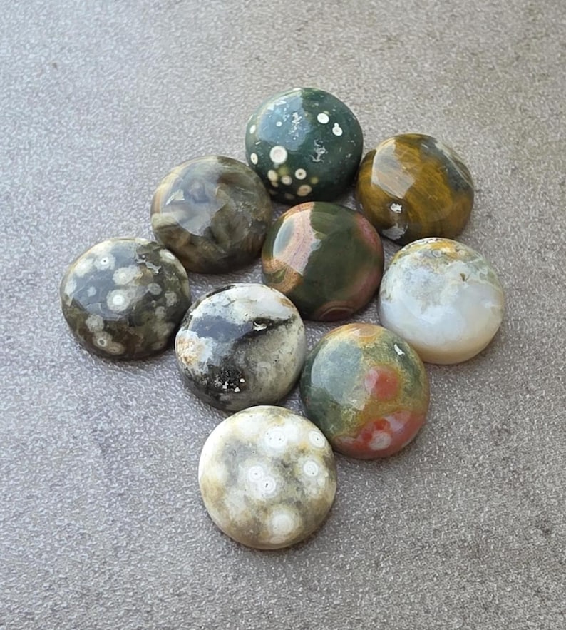 May include: A collection of nine polished gemstones in various colors and patterns. The stones include shades of green, brown, white, and red, with speckled and marbled textures. The stones are oval and round, and are arranged on a gray surface.