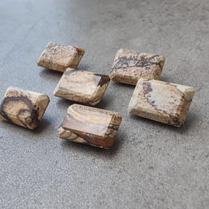以下が含まれることがあります： ベージュ、ブラウン、クリーム色の天然石で作られた、長方形のビーズが6つ。ビーズは面取りされた幾何学的な形状で、研磨された表面には石のユニークな模様が映し出されています。