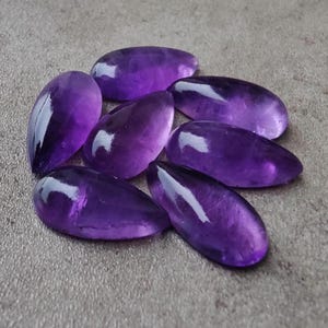 Op de afbeelding: Een groep van zeven amethist edelsteen cabochons in een traanvorm. De cabochons zijn diep paars van kleur en hebben een gladde, gepolijste afwerking.