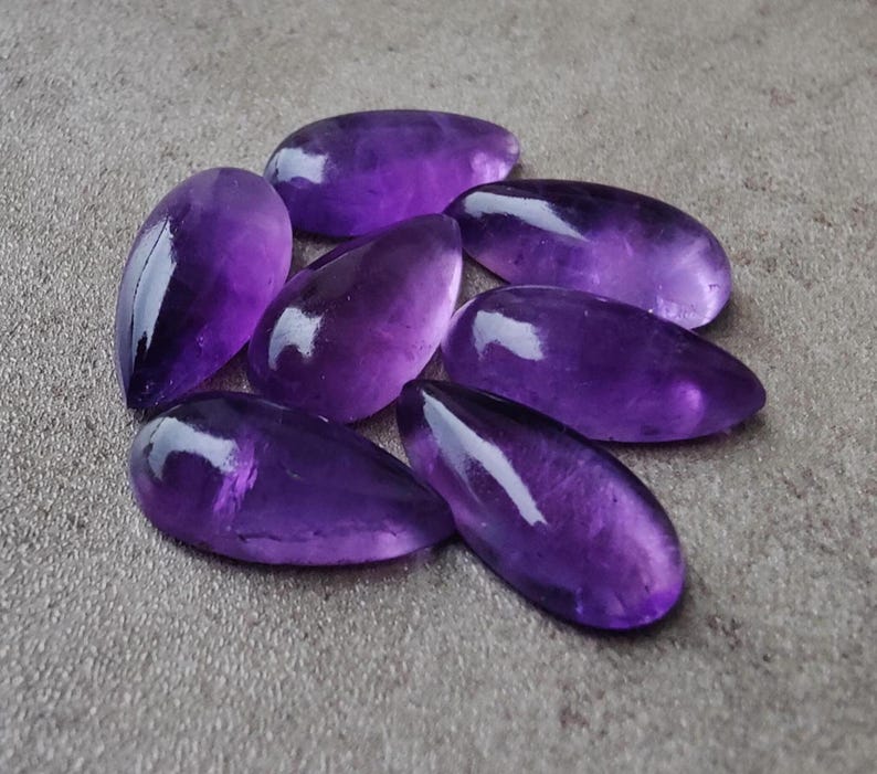 Op de afbeelding: Een groep van zeven amethist edelsteen cabochons in een traanvorm. De cabochons zijn diep paars van kleur en hebben een gladde, gepolijste afwerking.