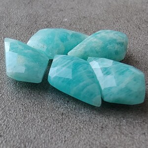 May include: Five teal green amazonite gemstone beads with faceted edges. The beads are arranged in a cluster.