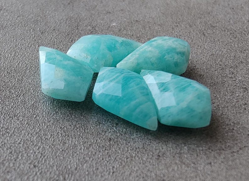 May include: Five teal green amazonite gemstone beads with faceted edges. The beads are arranged in a cluster.