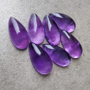 Op de afbeelding: Acht druppelvormige amethist cabochons in verschillende tinten paars. De cabochons zijn glad en gepolijst, met een glanzende afwerking.