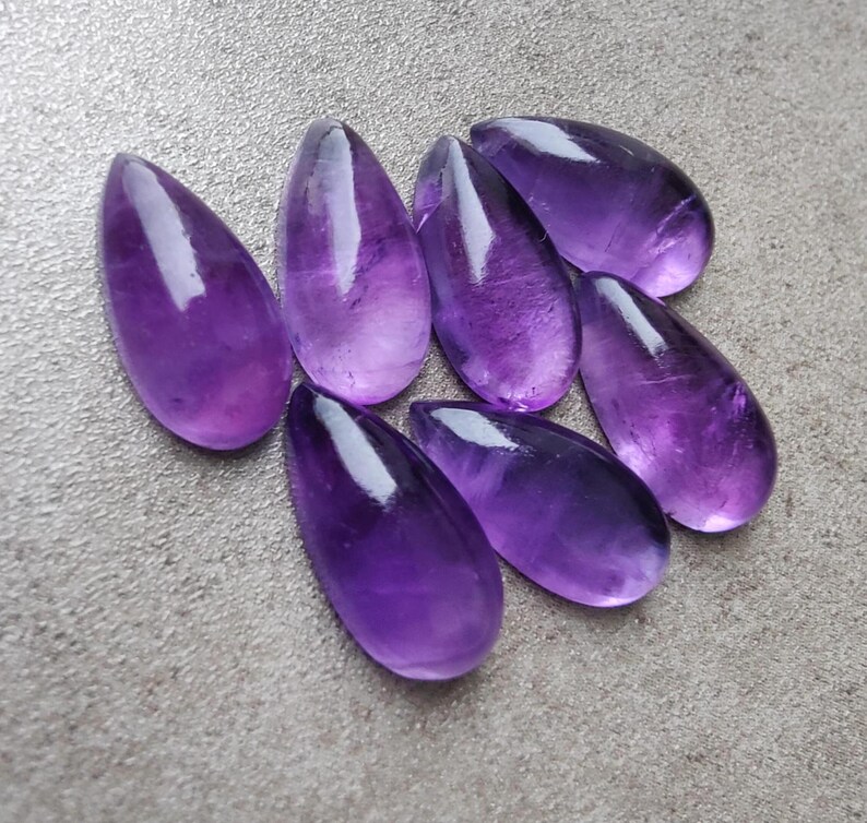Op de afbeelding: Acht druppelvormige amethist cabochons in verschillende tinten paars. De cabochons zijn glad en gepolijst, met een glanzende afwerking.