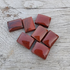 Puede incluir: Siete piedras de jaspe rojo lisas y pulidas, cada una con una forma y un tamaño ligeramente diferentes, dispuestas en un grupo sobre una superficie de madera clara.