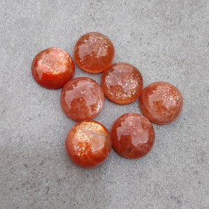 May include: Seven round, orange-brown sunstone cabochons with a shimmery, iridescent finish. The stones are arranged in a semi-circle on a gray surface.