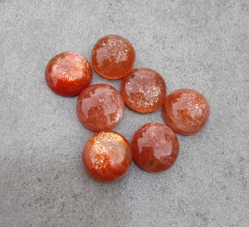 May include: Seven round, orange-brown sunstone cabochons with a shimmery, iridescent finish. The stones are arranged in a semi-circle on a gray surface.