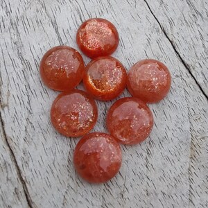 May include: Seven round, orange sunstone cabochons with a sparkly, iridescent finish. The stones are arranged in a loose cluster on a light wood surface.
