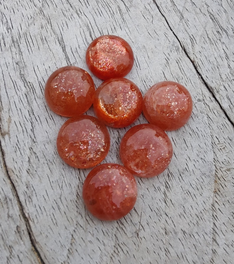 May include: Seven round, orange sunstone cabochons with a sparkly, iridescent finish. The stones are arranged in a loose cluster on a light wood surface.
