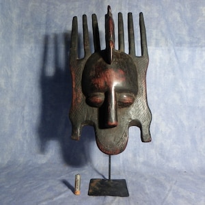 Puede incluir: Una máscara tribal de madera oscura con un rostro humano y una parte superior en forma de peine. La máscara está montada en un soporte de metal negro.