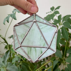May include: A geometric stained glass suncatcher in a dodecahedron shape. The glass is a pale green with darker green veining, framed by silver-colored metal. The suncatcher is suspended by a thin wire, and is displayed against a backdrop of green foliage.