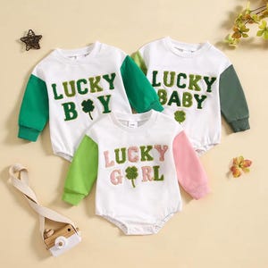 May include: Three white baby rompers with long sleeves. Each romper has a different color sleeve and the words "LUCKY BOY", "LUCKY BABY", and "LUCKY GIRL" embroidered on the front, along with a shamrock.