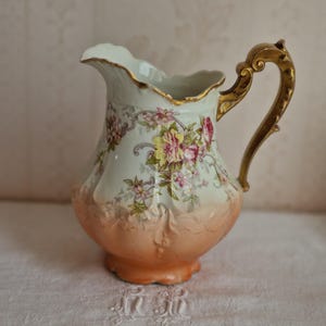 Peut inclure: Un pichet en porcelaine avec un corps supérieur crème orné de motifs floraux roses et jaunes, un bord festonné avec une bordure dorée, une base pêche avec des volutes en relief et une anse dorée incurvée et ornementée.