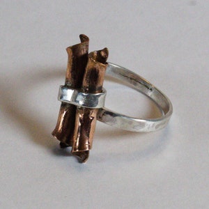 May include: A handcrafted ring featuring a silver band and a cluster of copper-colored, curled metal elements. The ring's design incorporates a silver band that wraps around the copper elements, creating a unique and artistic piece of jewelry.