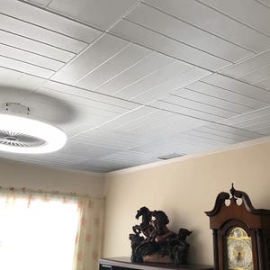 May include: A white ceiling with a grid pattern of rectangular panels. A white ceiling fan with a light fixture is in the center of the image. The ceiling is in a room with beige walls and a window with white curtains.